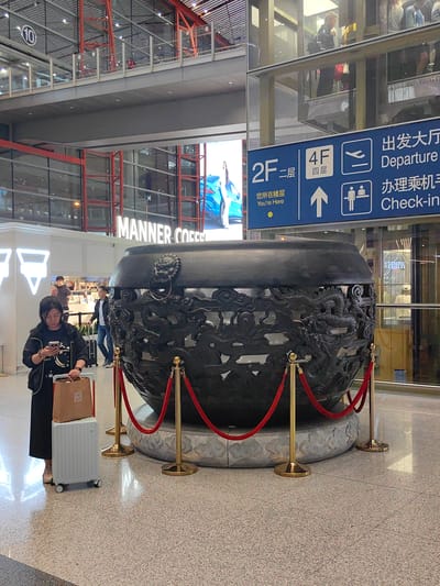The image shows an interior shot of an airport, likely in the city of Shunyi District, China.

A woman is seen standing to the left of a large, ornate, dark-colored decorative pot, with a suitcase beside her and a shopping bag resting on top of the suitcase. She appears to be looking at her mobile phone. Behind her, two men are walking, one with a suitcase.

The pot is encircled by red velvet ropes held by gold stanchions. Behind the pot is a glass-enclosed elevator. In the background, there's a "MANNER COFFEE" store and an advertisement featuring a blue car.

Above the store, there is an airport directory sign showing floors 2F and 4F. The sign includes the words "Departure" and "Check-in" in English and Chinese. The number "10" in a circle can be seen near the ceiling.

The floor is a speckled, light-colored tile. The building's architecture includes a mix of exposed metal structures and glass walls.