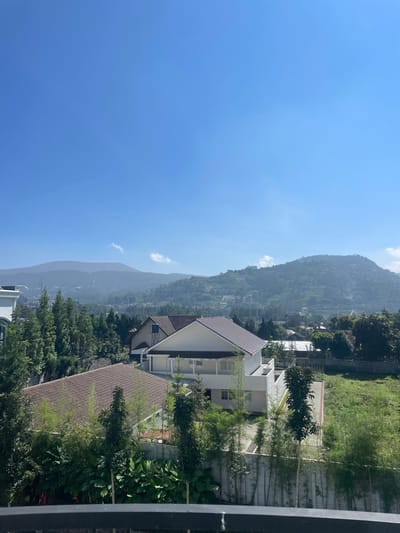 This image, captured in Kayuambon, Indonesia, presents an elevated daytime landscape view. The foreground features the upper section of a dark, metallic railing, indicating the vantage point is from a balcony or similar structure. Below the railing, dense green vegetation dominates, including large-leafed plants and several tall, slender trees, some resembling bamboo.

In the midground, residential structures with distinct architectural features are visible. A prominent, two-story house with white walls and a brown tiled roof occupies the central-right. Another house with a similar roof is partially visible to its left, and a larger white building is on the far left. The houses are surrounded by a variety of trees and shrubs, including coniferous species. A bare or sparsely vegetated plot of land is visible to the right of the central house.

The background is dominated by multiple layers of hills and mountains. These landforms exhibit diverse vegetation, ranging from dense forests to what appear to be terraced agricultural fields on some slopes. A light atmospheric haze partially obscures the distant mountains. The sky above is clear and uniformly blue, with a few wispy white clouds visible near the mountain peaks on the right side. No people or animals are discernible in the scene.