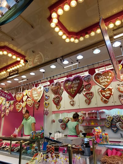 The image shows a stall at a fairground in Cottbus - Chóśebuz, Germany. The stall is decorated with numerous heart-shaped gingerbread cookies, many of which are adorned with phrases like "Ich liebe dich" (I love you). The cookies are hung from a rack above the counter. In the background, a woman with her back to the camera is serving customers. In the foreground, a man in a green polo shirt is standing behind the counter, and a display case filled with various candies and sweets is visible. The stall has a pink backdrop and is illuminated by overhead lights and strings of decorative bulbs. The overall scene depicts a festive atmosphere common at local fairs.