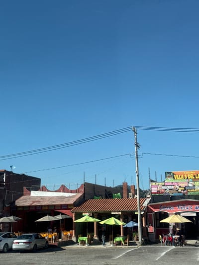 A vibrant street scene features restaurants with outdoor seating under colorful umbrellas. Overhead, power lines stretch across a clear blue sky. Signage for "Cabaña Villa's del La" and "Cabaña Villa's de" is visible.  The restaurant offers desayunos (breakfast), huaraches, chorizo, pambazo, and other dishes. Several cars are parked along the side of the street.