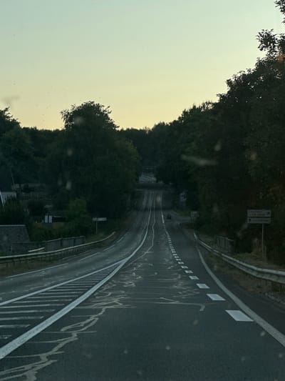 The image shows a road in Gruchet-le-Valasse, France, at dusk. The road is asphalt with white markings and a guardrail on the right side. Trees line the sides of the road, and a sign indicating directions to Gruchet-le-Valasse and Lillebonne is visible on the right. The sky is clear with a gradient of light blue to yellow. There are a few cars on the road, and a small building is visible on the left side of the road. The overall scene is calm and peaceful.