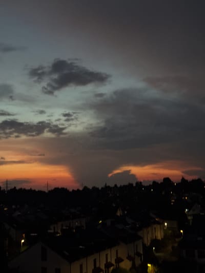 A high-angle, long-exposure photograph captures a twilight sky over a residential area in Sindelfingen, Germany. The sky is a gradient of colors, transitioning from a pale blue-gray at the top to hues of orange and pink near the horizon, indicative of a sunset or sunrise. Dark, scattered clouds populate the sky. Below the sky, silhouetted trees form a dark line against the illuminated horizon. The foreground is dominated by a dense cluster of residential buildings, appearing dark and indistinct due to the low light conditions. Small, scattered lights emanate from some of the buildings, suggesting inhabited homes. The overall atmosphere is calm and quiet.