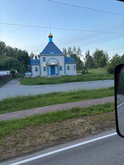 The image shows a small, light-colored church with a blue roof and a golden dome, located in the city of Kostrovo, Russia. The church has arched windows and doors, and is surrounded by green grass and trees. There are several blue bollards lined up on the asphalt road in front of the church. The sky is clear and blue. The photo appears to have been taken from a moving vehicle, as evidenced by the blurred foreground and the presence of a side-view mirror in the right side of the frame.