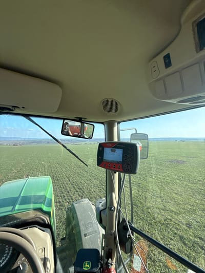 The scene depicts a view from the interior of a John Deere tractor cabin, looking forward over an expansive green agricultural field in Gagovo, Bulgaria. The sky is clear and blue, extending to a distant horizon where faint outlines of land or structures are visible.
Inside the cabin, an operator is partially reflected in the central rearview mirror, holding a mobile phone with a red/orange case, suggesting they are capturing the image. The tractor's steering wheel is visible in the lower left, along with a portion of the green hood. A black John Deere branded pouch rests near a small red fire extinguisher on the lower right. Mounted on a vertical pillar to the right of the windshield is a "KUHN SUNTRAK II" control monitor, displaying operational data and a graphic representing an agricultural implement. A single windshield wiper blade rests horizontally across the upper portion of the windshield. The cabin's interior features a beige headliner with a ventilation grille and an overhead console equipped with switches. Electrical cables are visible, connecting to the control monitor. A red/orange piece of equipment is reflected in the rearview mirror, situated behind the tractor.