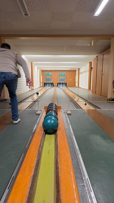 The image captures an indoor bowling alley, likely a "Kegelbahn" typical of Germany, with a person actively engaged in bowling. The perspective is from behind the bowler, looking down the length of two parallel lanes toward the pins.
In the foreground, a person, seen from the waist down and from behind, wears blue jeans and a light-colored long-sleeved top. Their right arm is extended forward, indicating they are either releasing or have just released a bowling ball. To the immediate right, a rack holds several bowling balls in dark blue-green and brown-maroon hues, waiting to be used.
The bowling lanes feature a distinct light green central track for the balls, bordered by prominent orange wooden rails that also function as ball returns. The main playing surface of the lanes is a grey-green material with fine horizontal grooves. Further down the right lane, a small dark bowling ball is visible. At the far end, two sets of bowling pins are arranged on the lanes, illuminated by overhead lighting. Above each set of pins, an electronic scoreboard displays glowing graphics, presumably showing pin layouts and scores, though specific text is unreadable.
The interior walls are light-colored, featuring sections with square panels and a pattern of small dark dots, as well as areas with horizontal perforations. The space is brightly lit by overhead fluorescent lights. Wooden doors and paneling are visible at the far end of the alley, with some indistinct green illuminated elements on the doors, possibly indicators or signage.
The scene depicts a recreational activity within a functional and well-maintained facility in Guxhagen, Germany.