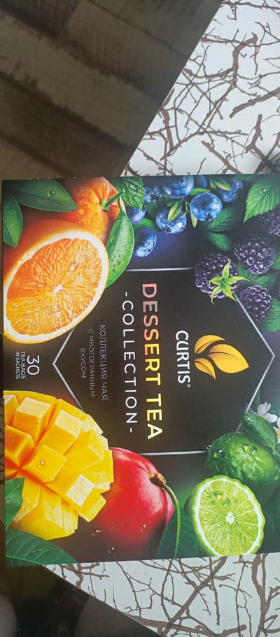 A box of Curtis Dessert Tea Collection is displayed on a wooden surface. The box features images of various fruits, including oranges, blueberries, blackberries, mango, and lime. The text on the box is partially in English and partially in Russian. The English text reads "CURTIS DESSERT TEA COLLECTION" and "30 TEA BAGS IN SACHETS". The Russian text reads "КОЛЛЕКЦИЯ ЧАЯ С МНОГОГРАННЫМ ВКУСОМ" which translates to "TEA COLLECTION WITH A MULTIFACETED TASTE". The box is positioned at an angle, and the wooden surface appears to be a table or floor. The background includes a white surface with a brown, branch-like pattern. There are no people or actions depicted in the image. The location of Sokol, Russia, is not discernible from the provided image.