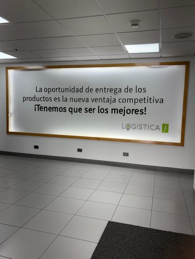 The image shows a brightly lit indoor scene, likely a commercial or office space, with a white tiled floor and a drop ceiling. A large, illuminated sign with a wooden frame is mounted on a white wall. The sign displays text in Spanish and a logo. The text reads: "La oportunidad de entrega de los productos es la nueva ventaja competitiva iTenemos que ser los mejores!" which translates to "The opportunity for delivery of products is the new competitive advantage We have to be the best!". To the right of the text is a logo that includes the word "LOGISTICA" with a stylized green 'f' and a map pin icon. The overall impression is one of a modern business environment emphasizing efficiency and competitiveness. There are no people visible in the image. The lighting suggests it could be daytime, but the indoor setting makes this uncertain. The context provided, Villa El Salvador, Peru, indicates the geographical location of this business.