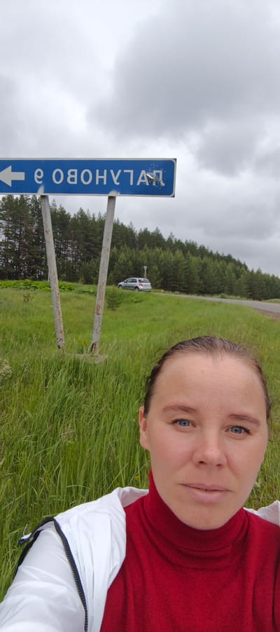 An adult female is depicted in a selfie, positioned in the foreground of a rural outdoor scene in Makshaki, Russia. She has light skin, blue eyes, and light brown hair pulled back. She is wearing a red turtleneck sweater under a white zippered jacket and is looking directly at the camera. Behind her, a blue rectangular road sign with white mirrored Cyrillic text, which appears as "ЯТЛАНУВО О" and includes a left-pointing arrow, is mounted on two weathered metal poles. In the middle ground, a silver SUV is visible on a paved road that curves into a dense evergreen forest. The immediate foreground consists of tall, verdant green grass and other foliage. The sky above is overcast with light gray clouds.