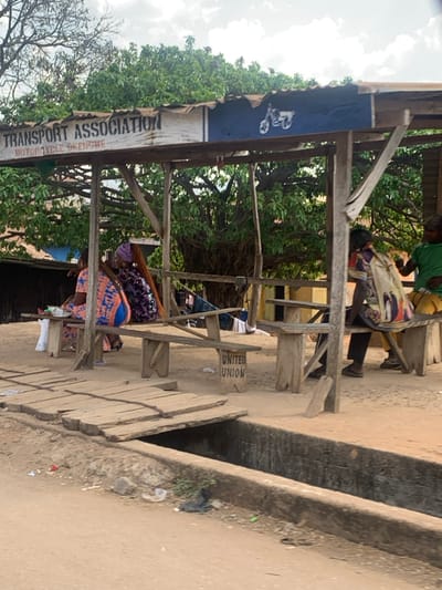 This image depicts a roadside scene in Egbedi, Nigeria, focusing on a rudimentary open-sided shelter identified as a "TRANSPORT ASSOCIATION MOTORCYCLE BKENGBWE" station. The shelter features a corrugated metal roof supported by rough-hewn wooden posts and beams. A banner on the roof displays the aforementioned text and a white motorcycle silhouette on a blue background.
Inside the shelter, five individuals are present, seated or resting on simple wooden benches and tables. From left to right:
1. A person in an orange and blue patterned dress is seated on a bench, facing away. A white bag rests beside them.
2. Another person, wearing a purple patterned outfit and a purple headwrap with long reddish-brown hair, is also seated on a bench, facing away.
3. In the mid-ground, a less distinct figure appears to be reclining or draped over a bench. Clothes are visible, possibly drying or stored, over a wooden beam. A sign on the ground reads "UNITED UNION".
4. A fourth individual, wearing a light-colored patterned tunic, is seated with their back partially to the camera, a colorful patterned bag resting near them.
5. The fifth person, wearing a green shirt and yellow trousers, sits further right, facing partially away from the camera.
The ground around and within the shelter is unpaved, appearing as dirt or sand. A concrete drainage ditch runs horizontally across the foreground, separating the shelter from the viewer. A makeshift wooden bridge, constructed from planks, spans this ditch, providing access to the shelter. Scattered debris, including plastic bags, is visible on the unpaved ground adjacent to the ditch. The background features dense green foliage from trees, under an overcast sky.