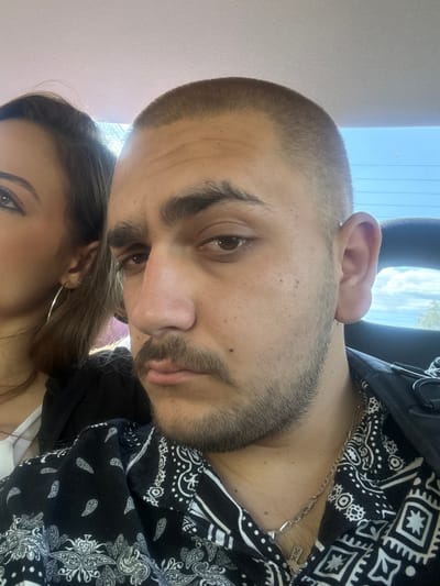 A close-up, rotated, tilted photo depicts a man and a woman in what appears to be a car. The man, whose face is the primary subject, has short, cropped hair and a full beard. He is wearing a black and white patterned shirt, possibly a bandana print, and a silver chain is visible around his neck. The woman is partially visible in the lower portion of the frame. Only part of her face, including an eye and her ear, is visible. She has long brown hair and is wearing a hoop earring. Light is coming from the top left. The location in the city of Zlatna Panega, Bulgaria, cannot be determined from the photo.