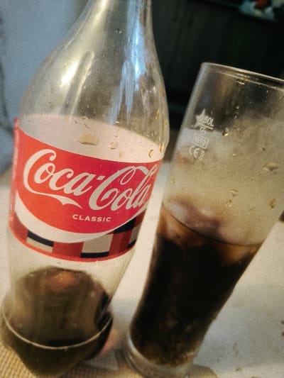 In a close-up shot, a partially empty bottle of Coca-Cola Classic stands next to a glass of the beverage. The bottle, made of clear plastic, has condensation on its surface. The iconic red and white Coca-Cola logo is prominently displayed on a label, with "CLASSIC" printed below the brand name. The lower part of the bottle appears to contain some of the dark brown liquid.
To the right, a glass, also exhibiting condensation, is filled with Coca-Cola. The dark liquid is visible in the lower portion of the glass, and some foam or bubbles are present near the top. Etched into the glass are markings, including "0.50L," "M19," and "CE," indicating its capacity and manufacturing details.
The scene is set on a textured surface, possibly a table, with a light-colored tablecloth or placemat. The background is blurred, suggesting an indoor setting with a muted, possibly dark, environment. The lighting appears to be artificial, casting highlights on the bottle and glass. The overall mood is casual, perhaps depicting a moment of refreshment or a pause during a meal. The image's grainy texture adds a vintage or artistic quality to the composition. There are no people visible in the image, only the objects and their immediate surroundings. The location context, Асқар Тоқпанов ауылы, Kazakhstan, suggests a rural or village setting in Kazakhstan. The time of day and weather conditions are not discernible from the image itself.