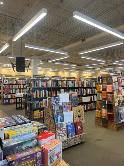 This image depicts the interior of a large retail bookstore located in El Paso, United States, consistent with the architectural and merchandising style of a Barnes & Noble establishment.
The environment features a high, industrial-style ceiling with exposed metal decking, structural trusses, and beige cylindrical HVAC ducts. Numerous bright white, rectangular linear LED light fixtures are suspended from the ceiling at various heights, providing general illumination. The floor is covered with a dark green or brown carpet.
The scene is dominated by multiple rows of dark wooden bookshelves, densely filled with thousands of books across various genres. Overhead signs, suspended from the ceiling, identify different departments, including "For Kids," "Art, Fashion & Photography," "Toys & Games," and "Gift Wrap."
In the foreground, a low display surface presents a collection of boxed items, likely children's games, crafts, and educational toys, such as "Sensory Squish Bag," "The Search for UAPs," and "Nature Crafts." Adjacent to this, a prominent, multi-tiered display stand with a patterned base showcases larger format books, including titles like "Linda Evangelista Steven Meisel," "Chagall," and "Ranchland." To the right, a tall, narrow display unit features an assortment of smaller bound items, such as journals or gift notebooks, in various colors.
Further into the store, a dark-colored table with stacked books is visible between bookshelves. Two dark, flat-panel monitors are mounted back-to-back on a central light-colored column on the left side of the frame. No individuals are visible within the bookstore.