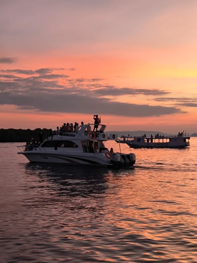 The image depicts a serene sunset scene over a body of water, likely in Rinca Village, Indonesia. Two boats are the primary objects, both carrying groups of people. The foreground features a white motorboat with "Hope" written on its side, its stern equipped with two large outboard motors. Several individuals are visible on its deck, some standing and observing the surroundings, with one person appearing to take a photograph. A life preserver is also visible on the boat.
In the background, a larger, elongated vessel, possibly a ferry or a tour boat, sails on the water. This boat also carries a substantial number of passengers, many of whom are silhouetted against the colorful sky. The boats are navigating calm waters, reflecting the warm hues of the sunset.
The sky is a prominent feature, painted with soft gradients of pink, orange, and yellow, indicative of either sunrise or sunset. A few scattered clouds add texture to the sky. Distant land, appearing as a dark silhouette of trees and rolling hills, forms the horizon.
The overall mood is peaceful and idyllic, suggesting a leisurely evening activity, possibly a sunset cruise or a journey back to shore. The warm colors of the sky and water evoke a sense of tranquility and natural beauty. The presence of people enjoying the view on the boats suggests a shared experience of appreciating the natural spectacle.