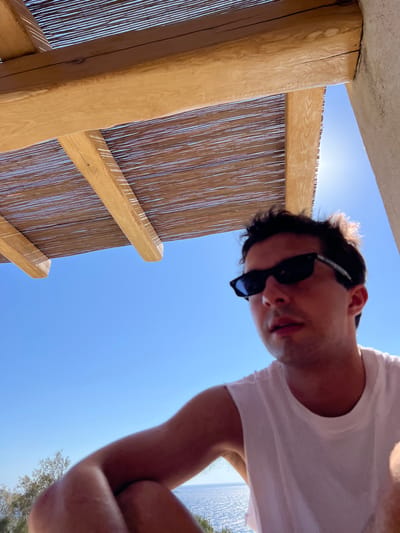 The image displays a male subject situated outdoors under a bright sky in Agios Ioannis Diakoftis, Greece. The individual, positioned in the lower right and center of the frame, wears a white sleeveless shirt and black framed sunglasses. His head is slightly turned to his right, and his mouth is partially open. Above the subject, a wooden pergola with a woven reed or bamboo matting roof is prominently featured, casting striped shadows. Intense sunlight emanates from the upper right quadrant of the image, creating a bright glare. The background reveals a clear, vibrant blue sky transitioning to a sparkling blue body of water, indicative of the Aegean Sea. Sparse green foliage is visible on the left side of the frame, and a section of a light-colored, possibly stucco, wall or column is present on the far right. The subject appears to be in a relaxed, seated or semi-squatting posture, looking out towards the distant water.