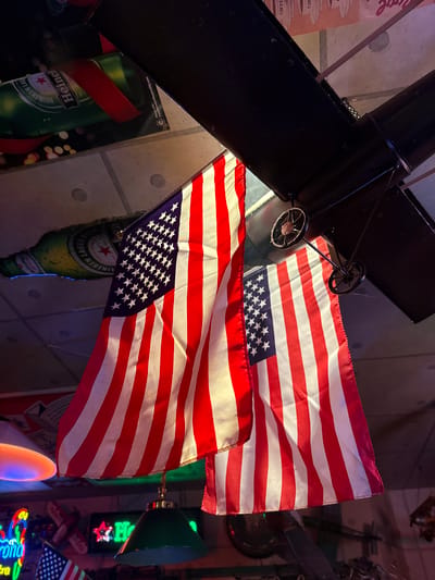 Two American flags are suspended from the ceiling in what appears to be a bar or restaurant. One flag is larger and positioned more prominently in the foreground, while a second, slightly smaller flag hangs behind and to the right of it. Both flags are draped and partially obscuring other elements of the scene.
The ceiling is made of concrete tiles and appears to be industrial in style. There are also elements of decor visible, including what looks like a model airplane hanging from the ceiling with visible wheels and struts. Behind the flags, the ceiling features promotional material for Heineken beer bottles. One bottle is particularly prominent, depicted in a green bottle with red and white labeling, wrapped with a red ribbon.
Below the flags, the environment becomes more cluttered and indicative of a commercial establishment. A green pendant light fixture hangs from the ceiling. Further in the background, neon signs are visible. One appears to be for "Corona" and is lit with colorful lights, while another green neon sign with a red star and "Heineken" lettering is also present. Other indistinct objects and lighting effects suggest a dimly lit, potentially lively atmosphere. The overall impression is of a themed bar or entertainment venue decorated with American and beer-related memorabilia.
This scene is not in Adachi, Japan.