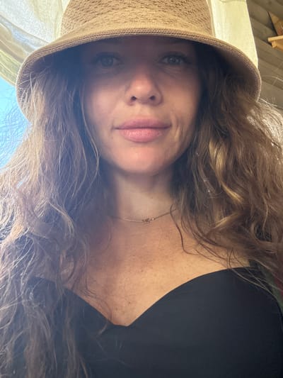 A woman with long, wavy brown hair and blue eyes, wearing a straw hat and a black off-the-shoulder top, is pictured from the chest up. She is looking directly at the camera and has on light pink lipstick and a thin gold necklace. The background shows a light-colored curtain, blue sky, and wooden structures, suggestive of an outdoor or partially covered setting. The location is Kupchegen, Russia.