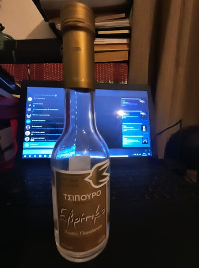 The image shows a glass bottle of Tsipouro, a Greek alcoholic beverage, placed in front of an open laptop. The bottle has a gold-colored cap and a label with Greek text. The laptop screen displays various chat windows and a background with a blueish hue. Books are stacked behind the bottle and laptop, and a curtain is visible in the background. The scene appears to be indoors, possibly in a room or office. There is no indication of the city of Xanthi, Greece in the image.