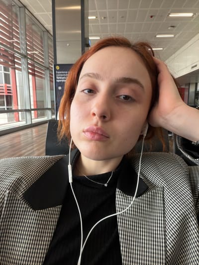 A female individual with reddish-brown hair is depicted in a close-up, high-angle selfie, looking directly at the camera with a neutral expression and slightly pursed lips. She is wearing wired white earbuds, a black ribbed top, and a black and white checked blazer. A silver chain necklace with a small circular pendant is visible around her neck. Her left hand rests near the left side of her head. The background indicates an indoor waiting area within a modern airport, specifically Sabiha Gökçen International Airport (SAW), which serves the Istanbul metropolitan area including the nearby district of Tuzla, Turkey. This is evidenced by large floor-to-ceiling windows revealing exterior red horizontal architectural elements, a prominent vertical metal pillar, and a partially visible yellow sign with black text advertising "Experience connectivity Türkiye's leading operator." Overhead grid lighting is visible on the ceiling.