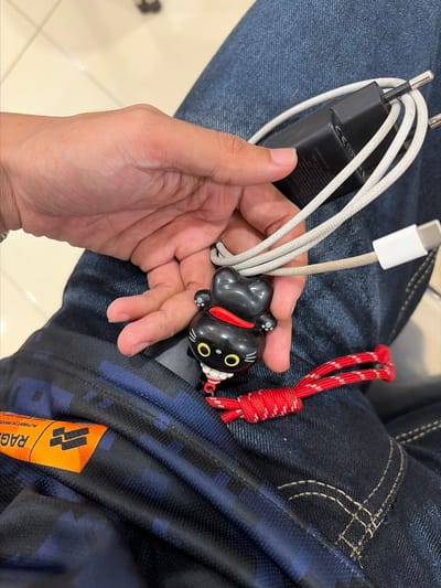 The image shows a close-up of a person's hand holding a black and white USB-C charging cable and adapter. Attached to the cable is a small, black, cat-shaped charm with yellow eyes and a red collar. A red braided cord is also visible, attached to the charm. The background consists of dark blue denim fabric, likely jeans, and a glimpse of a patterned blue and black fabric. Visible text on the adapter includes "UGREEN", "CE", "100-240V~50/60Hz 1.0A", "Output Power: 30.0W Max", and manufacturing information. A tag with the word "RAGE" is visible on the patterned fabric. The setting appears to be indoors, with a neutral-colored floor in the lower left corner. The lighting suggests it's daytime. The activity appears to be a person showing off or holding their charging accessories, with the decorative charm adding a personal touch. The focus is on the objects and the hand holding them.