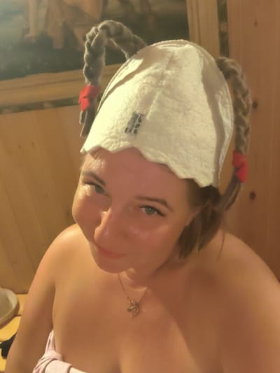 A woman, appearing to be in her late 20s to early 30s, is pictured in a wooden sauna. She is wearing a white felt sauna hat with two braided pigtails adorned with red ribbons. The woman is looking at the camera with a slight smile, and her shoulders and upper chest are visible, wrapped in a pink towel. She is wearing a silver necklace with a pendant. The background consists of warm-toned wooden walls, typical of a sauna interior, and a blurred image of a painting or tapestry on the upper left. The overall lighting is warm and dim, indicative of the sauna environment. The location is identified as Vostets SNT, Russia.