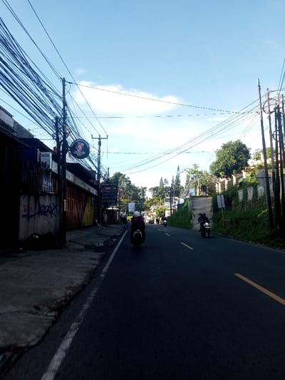This image captures a daytime street scene in Khouathineung, Laos, under a clear blue sky with sparse clouds. The asphalt road, marked with a solid white line on the left shoulder and a double solid yellow line indicating the center, extends into the distance, appearing to curve and ascend slightly.
On the left side of the road, a concrete sidewalk borders a series of commercial buildings, some with closed metal roll-down shutters. Graffiti is visible on the lower section of one building's wall. Multiple wooden utility poles stand along the sidewalk, supporting a dense tangle of overhead electrical and telecommunication wires that crisscross the frame. A circular sign, featuring a light-colored character on a dark background, is affixed to one of these poles, alongside another rectangular sign further down the street.
The right side of the road features a rising, vegetated embankment covered in green foliage, including shrubs and trees. A white, possibly stone, wall runs along the upper part of this embankment. Additional utility poles with wires are also present on this side, and a concrete stairway is visible ascending the hillside further along the road.
Three individuals are visible on motorcycles on the road. In the foreground, a single motorcyclist wearing a white helmet and dark attire rides a dark-colored scooter away from the viewer. Further ahead in the same lane, a second motorcycle carries two individuals; the driver wears a dark helmet and blue top. A third motorcycle is partially visible in the distance on the right side of the road. No other vehicles or pedestrians are clearly discernible.