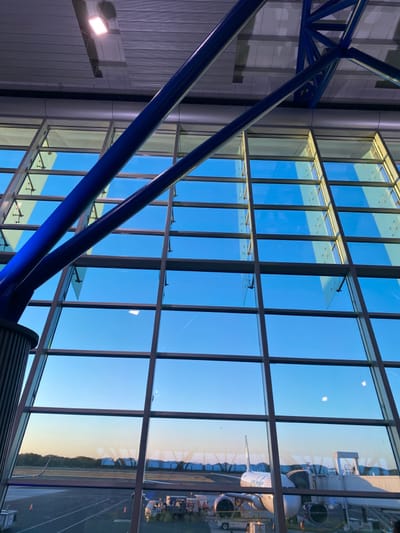 The image displays an interior perspective from Monseñor Óscar Arnulfo Romero International Airport in San Luis Talpa, El Salvador, looking through a large, gridded glass wall onto the tarmac. The scene is set under a clear blue sky, which lightens towards the horizon, indicating either early morning or late afternoon. In the foreground, two prominent diagonal blue metallic structural beams traverse the frame, originating from the upper left. A vertical fluted column is partially visible on the far left. The glass wall comprises numerous rectangular panes within a metal frame.

Beyond the glass, a white passenger aircraft is parked on the tarmac, occupying the lower right quadrant of the exterior view. A white passenger boarding bridge extends from the terminal, connecting to the aircraft's fuselage. Several ground support vehicles, including what appears to be a white fuel truck and other smaller service carts, are positioned near the aircraft's engines and undercarriage. Multiple individuals, identifiable as ground crew, are present on the tarmac in proximity to the servicing equipment and aircraft. The tarmac surface is dark and marked with white operational lines. In the distant background, a strip of low-lying green vegetation is visible beneath a distinct, patterned barrier structure, which horizontally spans the mid-ground of the exterior scene.