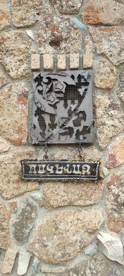 A weathered metal plaque featuring an intricate cutout design of leaves and vines is mounted on a rough stone wall. Below this plaque, a smaller, rectangular sign with ornate gold and white lettering hangs by chains. The sign has a dark background and appears to be made of metal or wood with painted details. The lettering on the sign is in Armenian script. The stone wall itself is constructed from irregularly shaped, beige and brown stones of varying sizes, held together with gray mortar. Some of the stones have a slightly textured or mossy appearance. The scene suggests an outdoor setting, possibly a historical building or a decorative element in a public space within the city of Voskevaz, Armenia.