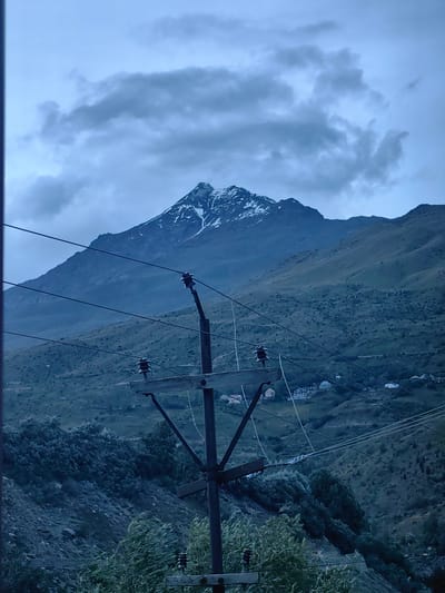 The image depicts a scenic mountain landscape under a cloudy sky, likely during dusk or dawn, given the muted light and cool tones. In the foreground, a utility pole with insulators and power lines cuts across the frame, obscuring parts of the background. The pole appears weathered and utilitarian, indicating human infrastructure within a natural setting.

The main subject is a majestic mountain peak, its upper reaches dusted with snow, contrasting with the green, undulating slopes below. These slopes are textured with vegetation, appearing somewhat barren in places and densely covered in others, suggesting a rugged terrain. Scattered across the hillsides are a few buildings, possibly a small village or isolated homes, hinting at human habitation within this remote location.

The weather appears overcast, with heavy, dark clouds dominating the sky. The lighting is dim, casting a somber mood over the scene. The overall atmosphere is one of quietude and natural grandeur, with a touch of human presence through the power lines and buildings. There are no people visible in the image, and no text is present. The location context provided is Gushal, India, which aligns with the mountainous terrain and potential for snow-capped peaks. The image captures a moment of serene, yet somewhat dramatic, natural beauty, juxtaposed with man-made utility elements.