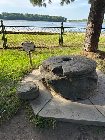 The image displays a large, circular millstone, possibly a historical artifact, placed on a stone base in an outdoor setting. To the left of the large millstone, a smaller, round stone object rests on the grass. A sign with Russian text, likely indicating the millstone's origin or historical significance, is positioned in front of the larger stone. The scene is situated near a body of water, likely a river, with a tree trunk visible to the right. The background shows a tree line and a blue sky. The location is identified as Terpilovo, Russia.