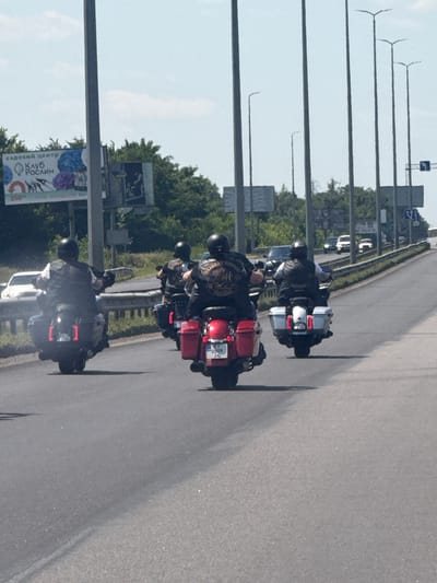 This image shows a group of motorcyclists riding on Peremohy Street in Ukraine. There are at least four motorcycles visible, each with a rider wearing a helmet and a vest. The motorcycles appear to be large cruiser-style bikes. The riders are traveling in the same direction on a paved road with other vehicles in the background. The environment is an urban or suburban setting, with trees and streetlights visible. The sky is clear and the lighting suggests it is daytime. The license plate on the second motorcycle from the left is partially visible and appears to read "KI 0009 JG".