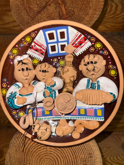A circular, decorative plate made of clay depicts a folk art scene. The plate is brown with an orange rim and is adorned with colorful, floral designs around the edge. In the center, three clay figures are arranged. On the left is a figure with a white headscarf, wearing a white shirt with blue and green trim. Next to it is a smaller figure, also in a white shirt with similar trim. On the right is another figure, playing an accordion, wearing a white shirt with blue and green trim. In front of the figures are various clay objects, including small pots and what appears to be a braided rope. Two small clay dogs are also present in the foreground. Behind the figures, there is a stylized depiction of a window with blue panes and a white and red embroidered cloth. The plate is displayed against a background of rough, natural wood. The scene is not located in Heiligenrode, Germany; it is a general folk art representation.