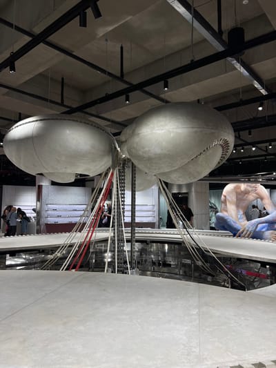 The image depicts an interior view of a modern commercial or exhibition space located in 성동구, South Korea. The environment features an industrial aesthetic with a raw concrete ceiling equipped with black track lighting.
Two large, light-grey, ovoid structures, appearing metallic or made of concrete, are suspended from the ceiling. The structure on the right exhibits a prominent textured opening, resembling a segmented shell or a mechanical gear. A complex central installation descends from between these suspended objects, consisting of multiple metal chains, white and red cables, and rods, converging towards a circular, light-grey platform. This platform forms a railing around a recessed, reflective lower level that mirrors the overhead structures.
To the left of the central installation, approximately five individuals are observed standing near illuminated white display shelves showcasing rows of eyeglasses. They appear to be browsing merchandise. One person in a black shirt stands closer to the central structure. To the right, a large, light-toned sculptural figure of a male torso, with significant portions of its lower body painted blue, is seated on an elevated platform. The background beyond includes further architectural and display elements.