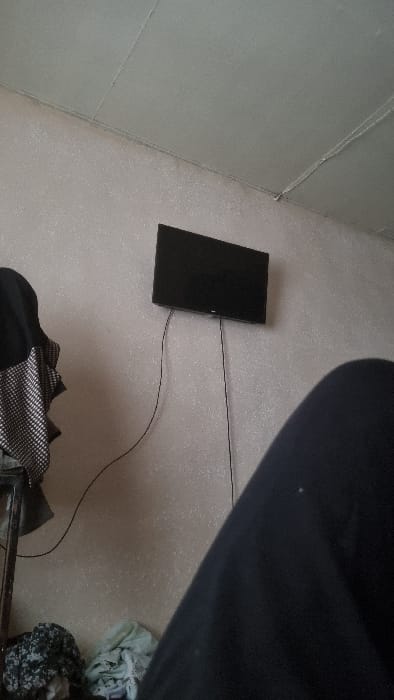 The image presents an overhead view of an indoor setting, taken from a slightly tilted perspective. The main surface visible is a light-colored floor, possibly carpeted or covered with a textured mat, which occupies most of the frame. On the left side, a light-colored wall or vertical panel is visible, with a seam running diagonally across it.
In the center-left of the image, a black rectangular electronic device, resembling a tablet or a large smartphone, lies flat on the floor. Its screen is dark, suggesting it is off or displaying a black image. A thin, black cable extends from the right side of this device, stretching towards the upper right and then curving down towards the bottom right of the frame.
In the upper right portion of the image, a large, dark, amorphous shape partially obscures the scene. This appears to be a portion of a person's leg or a dark piece of furniture, indicating the perspective of the shot.
Towards the bottom of the image, various items of clothing or fabric are visible. Immediately below the black device, there's a pile of dark fabric, including one piece with a distinct black and white checkerboard or patterned texture, and another plain black item. Further to the right and bottom, more crumpled light-colored fabric, possibly clothes, with a mottled or patterned design (resembling camouflage or a floral pattern), is seen on the floor. A metallic bar or frame, possibly part of a bed frame or chair, is visible along the very bottom edge of the image.
The lighting appears to be ambient indoor light. No specific activity is taking place, and the items are static. There is no visible text in the image. The overall impression is that of a casual, personal space, possibly a bedroom, where items are scattered on the floor.