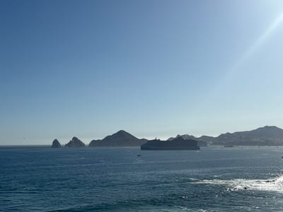 The image captures a coastal scene, likely in Cabo San Lucas, Mexico, given the distinctive rock formations in the distance. The foreground is dominated by the deep blue ocean water with moderate waves. A large cruise ship is anchored in the water, suggesting maritime activity. In the distance, land is visible, including rocky outcrops and coastal mountains. The sky is clear and blue, with the sun shining brightly in the upper right corner of the frame. The overall ambiance suggests a sunny day in a coastal tourist destination.