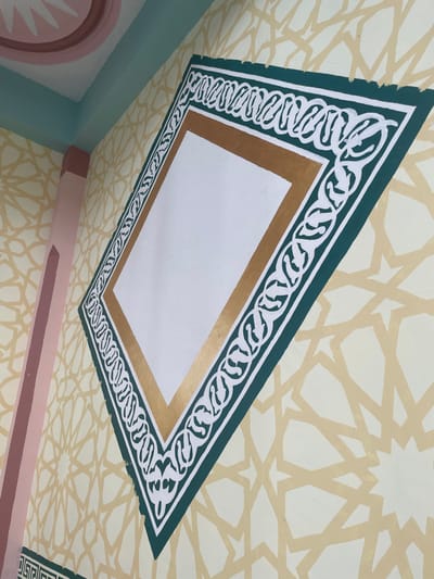 A brightly lit interior wall in Cutneuheun, Indonesia, displays decorative painting. A large diamond-shaped frame, rotated clockwise, is centrally positioned. This frame features a dark teal-green outer border with intricate white scrollwork, encasing a metallic gold inner border, which frames a solid white rectangular field. The wall background is light yellow-cream with a repeating, lighter beige Islamic geometric star pattern. To the immediate left, a peach-pink column extends vertically. Above, a light blue-green band separates the wall from a cream-colored ceiling, where a partial pink dome structure with a white starburst motif is visible in the upper left. No human presence or dynamic actions are depicted.