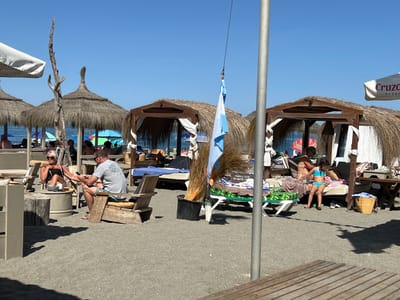 This media file captures a sunny day at a beach in San Roque, Spain, with several people relaxing under thatched-roof cabanas. The scene is set on a sandy beach, with numerous lounge chairs and umbrellas providing shade.
In the foreground, a man and a woman are seated on wooden chairs, engaged in conversation and looking at a smartphone. The woman is wearing a dark tank top and shorts, while the man is in a grey t-shirt and patterned shorts. A small table and a drink are visible between them.
Further back, more cabanas are visible, some with white curtains. People are lounging on sunbeds, some in swimwear. A young girl in a blue bikini is sitting on a sunbed, looking down. In the distance, the blue sky meets the calm sea.
A few notable objects include a black bucket filled with dried reeds, a blue and white flag flying from a string, and various beach umbrellas. One umbrella in the background displays the text "Cruzo" and "DESDE".
The bright sunlight and clear blue sky suggest it is midday. The overall atmosphere appears relaxed and leisurely, indicative of a vacation or a day spent enjoying the beach. The presence of the cabanas and lounge chairs points to a resort or a developed beach area.