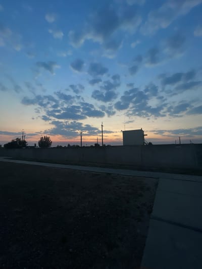 The image shows a vertical landscape of a sky at dusk, with some structures and trees visible on the right. The sky is filled with scattered clouds, tinged with hues of blue, gray, and soft orange, indicating either sunrise or sunset. In the middle ground, there is a low, rectangular building with a light-colored roof, and behind it, several utility poles with wires are visible. A line of trees and vegetation separates the structures from the foreground. The foreground on the right is a dark, textured area that appears to be rough ground or dirt, adjacent to a paved surface that could be a road or sidewalk. The overall mood is peaceful and serene, captured during twilight. There is no visible text in the image. The location is Mykolaivske, Ukraine.