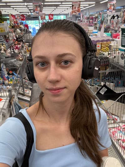 In a retail store in Erlenbach am Main, Germany, a young woman with blue eyes and brown hair pulled back in a ponytail is taking a selfie. She is wearing a light blue t-shirt and black headphones. The background shows aisles of merchandise, including toys, home goods, and clothing, as well as shopping carts. There are promotional signs with percentage symbols visible. The lighting suggests the store is well-lit with fluorescent ceiling lights.