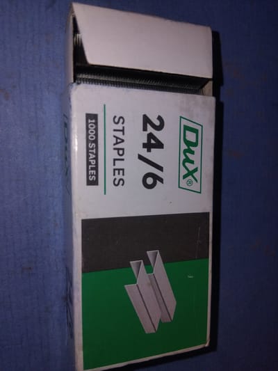 The image displays a box of Dux 24/6 staples, set against a blue textured surface, possibly a tablecloth or bedspread. The box is predominantly white and green, with the "Dux" logo prominently featured in green within a white rectangle. The number "24/6" is printed in large black font below the logo, indicating the staple size. Beneath this, "STAPLES" is written in smaller black font, followed by "1000 STAPLES" in a black rectangle with white text. On the right side of the box, a graphic illustration of two staples is visible on a green background. The staples themselves are partially visible on the left side of the box, showing a stack of metallic staples. The lighting in the image is somewhat dim, with a slight glare on the box. There are no people or animals present in the image. The scene appears to be an indoor setting, with the focus solely on the product packaging. The location context of Haveli Jhangranwali, Pakistan, is provided, but there are no specific visual cues within the image to confirm this location.