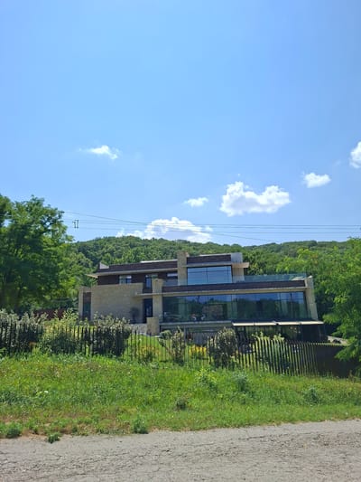The image shows a modern, multi-story house with large glass windows and stone facade, situated on a green hillside covered in trees. The house is surrounded by lush vegetation and a black metal fence. Power lines are visible in the sky, stretching across the frame. In the foreground, there is a rough, gray road with patches of grass. The sky is blue with scattered white clouds. The scene appears to be a sunny day in a rural or semi-rural setting. This property is identified as being located in Condrița, Moldova. There are no people or animals clearly visible within the frame, though some indistinct figures may be seen through the glass windows of the house.