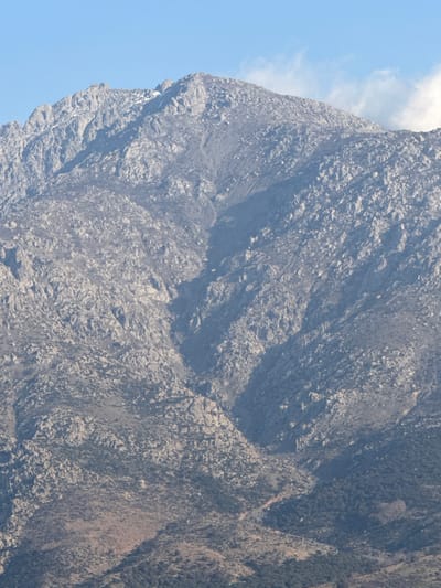 The image presents a rugged, rocky mountain under a clear blue sky with sparse white clouds on the upper right. This landscape is located in the vicinity of Kamariotissa, Greece. The mountain's upper sections feature exposed, jagged grey rock with small, isolated patches of snow or ice visible near the peaks on the left side. Descending slopes are characterized by extensive rockfall, scree, and interspersed areas of sparse, low-lying vegetation appearing as dark green or brown shrubbery. Several prominent, shadowed gullies traverse the mountain face from top to bottom. A faint, reddish-brown dirt path or track is discernible winding horizontally across the lower-middle portion of the mountain, cutting through the vegetation. Toward the bottom of the frame, the vegetation becomes denser, forming a more continuous dark green forest or scrubland. The scene is devoid of people, animals, or other man-made structures beyond the aforementioned path. Natural light illuminates the scene, creating distinct shadows on the right-facing slopes and within the gullies.