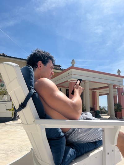 This low-angle image captures a man relaxing in a white reclining lounge chair under a bright, clear blue sky. The man, with dark, curly hair, appears to be bare-chested and is wearing dark blue trousers or shorts, resting on a dark blue cushion. He holds a black smartphone with a clear case in his right hand, displaying a gold-colored bracelet on his wrist. His head is tilted back, and he has a relaxed expression with a slight smile, looking upwards.

In the background, a stately building with a light-colored facade, reddish-pink trim, and prominent white columns is visible. Decorative finials, possibly resembling pineapples, are positioned along the roofline. Balconies with dark railings and potted palm trees add to the architectural detail. The sky is largely clear, dotted with a few wispy clouds and a distinct contrail.

The activity taking place is leisurely relaxation, with the man using his phone in an outdoor setting. The bright sunlight indicates it is daytime, likely midday or early afternoon, under sunny weather conditions. The architectural style and general atmosphere align with a Mediterranean resort or villa, consistent with the specified location of Èze, France. The overall impression is one of peace and enjoyment. No visible text is discernible in the image.