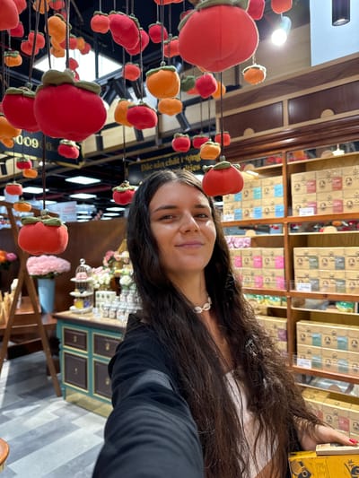 A woman is taking a selfie in a brightly lit indoor market or store. She has long, dark, wavy hair and is wearing a white top and a black jacket. She is also wearing a seashell necklace. In the background, there are shelves stocked with various items, many of which are in yellow and brown boxes. Several decorative, plush fruit ornaments, resembling persimmons, are hanging from the ceiling. The woman is holding a yellow box in her right hand. The scene suggests a commercial environment, possibly a gift shop or a specialty food store in Phu Thuy, Vietnam, given the presence of Vietnamese text on some of the items in the background.