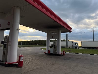 This is a full, eye-level, outdoor shot of a gas station on a cloudy day, possibly at dusk, with the sun breaking through the clouds on the left. In the foreground, to the left, is a white pillar supporting a red-trimmed canopy. To the right of the pillar is a gas pump. A red fire extinguisher on wheels is visible next to the pump. The ground is paved with hexagonal interlocking concrete pavers. Beyond the gas pump area, a road curves to the left, bordered by grass and a line of trees in the distance. Two white semi-trailer trucks are parked on the right side of the road. One truck has its cab visible. Overhead wires and a utility pole are visible in the sky. The scene is likely in Russkie Kirmeni, Russia.