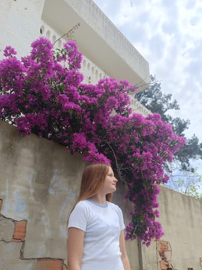 A young woman with long, reddish-blonde hair stands in front of a textured concrete wall adorned with vibrant purple bougainvillea. She is facing to her right, looking out of frame, with a calm and thoughtful expression. She wears a simple white t-shirt with a light blue trim around the neckline. The bougainvillea plant is lush and overflowing, with cascades of bright purple flowers spilling down the wall. Behind the flowers, a section of a white building with rounded decorative elements is visible. The sky above is overcast with soft, grey clouds. The wall itself shows signs of age, with patches of exposed brick and some blue graffiti marks. The overall scene suggests a warm day, possibly late morning or early afternoon, given the bright but diffused light. The setting appears to be outdoors in a Mediterranean climate, consistent with a location like Almardà, Spain, where such flowering plants are common.