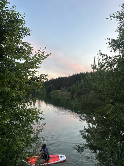 The image depicts a tranquil lake scene at dusk, framed by dense foliage on the left and right. In the foreground, a person wearing a blue life jacket is seated on a red and white paddleboard in the water, facing away from the camera. The lake surface is calm with gentle ripples, reflecting the soft hues of the twilight sky, which displays subtle shades of pink and pale blue. Across the lake, a dense forest of evergreen and deciduous trees lines the shore, with reeds visible along the water's edge. The overall atmosphere is serene and natural, suggesting a peaceful outdoor setting possibly in the vicinity of Gaiķi, Latvia.