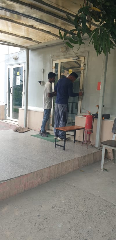 The image shows two men standing in front of a glass door at what appears to be an entrance to a building in Rooppur, Bangladesh. The man on the left is wearing a light-colored t-shirt and blue jeans. The man on the right is wearing a dark blue long-sleeved shirt and gray pants with black patches. He is seen interacting with the door mechanism, possibly attempting to unlock or repair it. To the right of the door, there is a red fire extinguisher mounted on a stand, a small wooden bench, and what appears to be a toolbox with a drill on top. The building has a covered walkway with a translucent ceiling and a light fixture visible. The ground in front of the entrance is paved with concrete tiles. There is some foliage from a tree hanging into the frame from the top right.