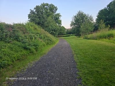 The image depicts a gravel path winding through a park-like setting under a clear sky. On the left side of the path, a dense, green hedge with scattered small pink flowers grows on an elevated bank. The right side of the path is a manicured lawn, dotted with small white flowers. Further into the park, several trees of varying sizes are visible. In the background, more trees and distant buildings can be seen. The timestamp in the lower left corner indicates "19 червня 2026 р. 9:00 PM," which translates to "June 19, 2026, 9:00 PM." This scene is located in Bannewitz, Germany.