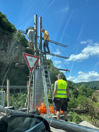 In the city of Dulovine, Montenegro, three men are engaged in construction work, possibly installing a signpost. One man, wearing a yellow shirt and grey trousers, is on a ladder, holding a tool. Another man, in grey shorts and a black t-shirt, is bent over a metal structure. The third man, wearing a bright yellow vest and dark shorts, stands facing away from the camera, observing the work. The background features a rocky hillside covered in lush green trees, under a bright blue sky with scattered white clouds. A red and white triangular yield sign is prominently displayed on a pole to the left of the construction site. Construction materials, including orange netting and large black pipes, are visible at the base of the structure. The overall scene suggests an active construction project in a scenic, mountainous area.