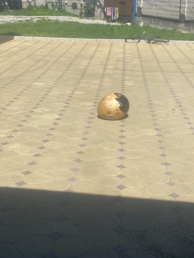 The image shows a paved courtyard with a patterned surface, likely in Belaya Rechka, Russia. A single, light-colored sphere with dark markings rests on the paving stones. To the right, a bicycle lies on its side. In the background, there is a patch of green grass, a building, and a clothesline with laundry. A shadow falls across the foreground of the image.